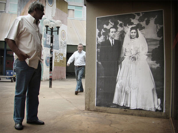 Foto horizontal no mesmo local anterior. A parede com a foto antiga ocupa metade da imagem. Há dois homens de meia-idade, um andando em direção à câmera, mais longe, e um parado no lado esquerdo da foto. Ele está de perfil, com as mãos no bolso, olhando para a foto antiga. Entre eles vê-se um poste de rua, preto e baixo, com três globos de vidro. 