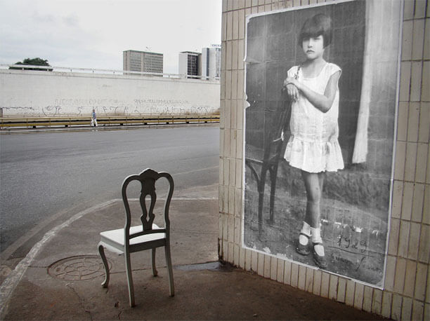 Foto horizontal mostrando a mesma foto antiga colada na parede, na metade direita da imagem. Na outra metade está uma cadeira de madeira de aparência antiga, cinza claro, virada para o fundo e para a esquerda. A rua está inteiramente vazia exceto por um homem bem ao longe, de pé, parado, encostado num guarda-corpo. Mais ao longe vê-se um paredão de concreto e o topo de três prédios.