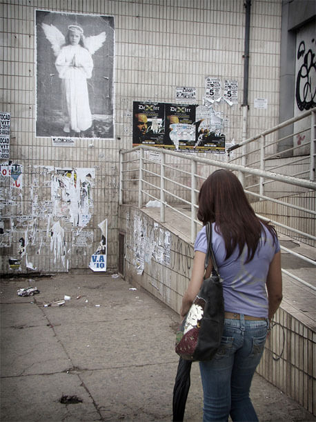 Foto vertical de uma mulher passando de costas para a câmera, à direita. Ela tem cabelos lisos e compridos, avermelhados; usa jeans e blusa lilás, carrega uma grande bolsa e um guarda-chuva. Está andando devagar, cabisbaixa, numa calçada de cimento. Do seu lado direito há rampa; à frente, uma parede alta que ocupa todo o fundo da foto. No canto esquerdo da parede, em cima, está colada uma grande foto vertical de uma menina em pé, de mãos postas, olhando para a câmera, com um vestido branco e longo de mangas compridas, cabelo louro comprido, asas e auréola de anjo. A parede do fundo e a da rampa são revestidas de ladrilhos antigos e muito sujos. Há vários cartazes colados, alguns sobre outros, em diferentes estados, alguns já totalmente arrancados. Nenhum foi colado em cima da foto antiga; eles respeitam uma certa distância e não passam da metade da sua altura, pois a foto está bem alta. 