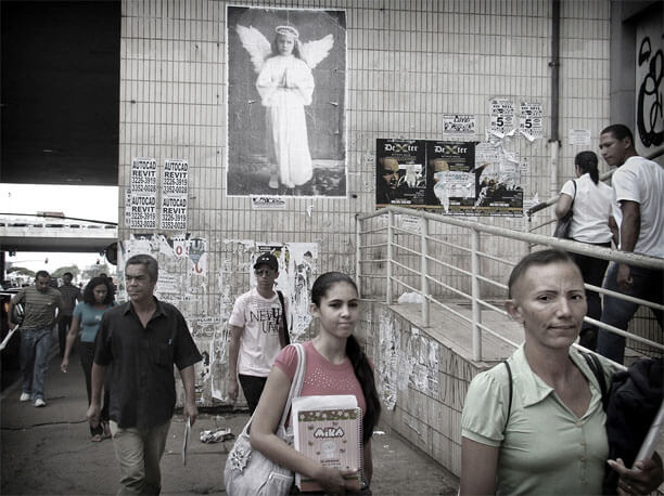 Foto horizontal do mesmo local anterior. No alto, ao centro, está a foto do "anjo". Do lado esquerdo do paredão vê-se um pedacinho da rua em perspectiva, o teto do viaduto próximo e outro viaduto ao longe. Há diversas pessoas andando em direção à câmera, algumas bem próximas e outras ao longe. A mais próxima, no canto direito, é uma mulher com a testa ligeiramente franzida, mas esboçando um sorriso e olhando para a câmera com um olhar intenso. Dois passos atrás dela há uma jovem que também olha para a câmera, com um meio-sorriso despreocupado.