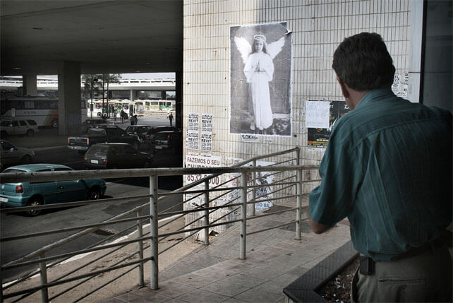 Foto horizontal do mesmo local anterior, mas num ângulo diferente. Foi tirada na entrada de um prédio, onde, no lado direito, um homem de costas se inclina para olhar para fora. A rampa de acesso ao prédio e seu guarda-corpo de ferro aparecem da esquerda para a direita. Na parte central, em cima, está a foto do "anjo" sobre a parede. À esquerda da parede vê-se a rua, carros passando, o teto do viaduto e da rodoviária, e ao longe alguns ônibus estacionados e pessoas andando. 