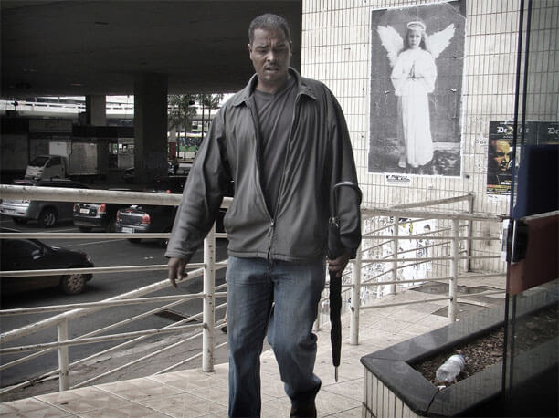 Foto horizontal no mesmo ângulo da imagem anteior. No centro da foto, bem perto, um homem de meia idade está chegando de frente, de cenho franzido e boca entreaberta, apressado, olhando para o chão. À direita dele vê-se a foto do "anjo" na parede. À esquerda estão a rampa, a rua com carros em movimento e a rodoviária.