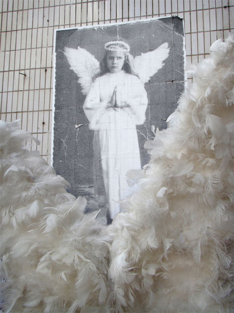 Foto vertical mostrando um detalhe de duas asas de uma fantasia de anjo. As asas são de penas brancas, em forma de V, e ocupam quase todo o quadro. Por trás do V aparece a foto do "anjo", dos joelhos para cima. A menina-anjo olha séria para a foto e pode-se ver melhor os detalhes, como as asas, a auréola e seu rosto expressivo.