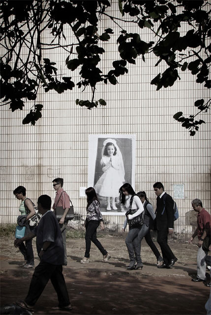 Foto vertical mostrando o paredão, o chão de terra e oito pessoas passando de perfil, da direita para a esquerda. Todas estão meio cabisbaixas. Do meio para a parte de cima da imagem vê-se as folhas de uma árvore, em silhueta, quase pretas. Na parte de baixo vê-se a sombra da árvore. Ao fundo, no centro, a foto antiga colada na parede.