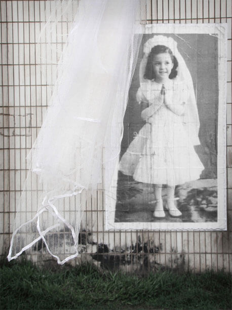 Foto vertical mostrando o detalhe da parte de baixo de um véu de filó branco. Ele sai da parte central, no alto, e vai até a parte de baixo, na esquerda. No fundo, à direita do véu e parcialmente encoberta por ele, vê-se a foto antiga mais de perto, com mais detalhes da menina e de suas roupas. Aparece uma pequena parte da parede degradada em volta da foto e do véu, e uma faixa de grama verde escura embaixo.