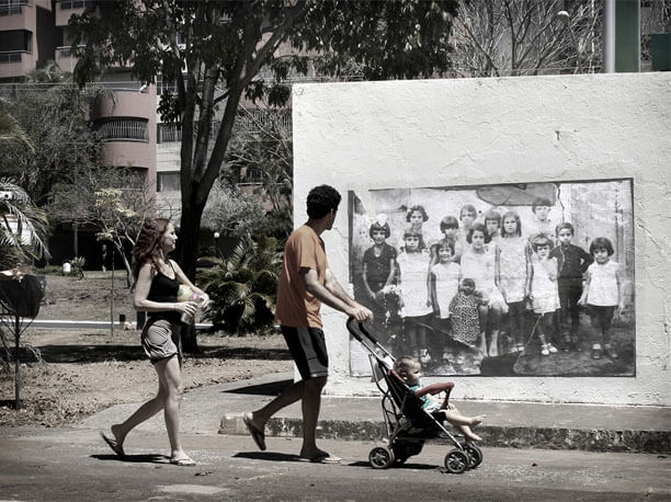 Foto horizontal de um casal passando com um bebê num carrinho, todos de perfil, olhando uma foto antiga colada na parede. A foto colada tem cerca de 3m x 2m e mostra um grupo de 14 crianças, que têm entre 4 e 11 anos de idade. As crianças posam olhando para a câmera. Todas estão de pé, menos a mais velha; ela está sentada no centro do grupo e segura pela mão uma bebê de dois anos ou menos, que está em pé, de vestido e touca. Na foto maior, atrás da parede, aparece um gramado, uma calçada, uma árvore, algumas plantas e um prédio de apartamentos ao fundo.