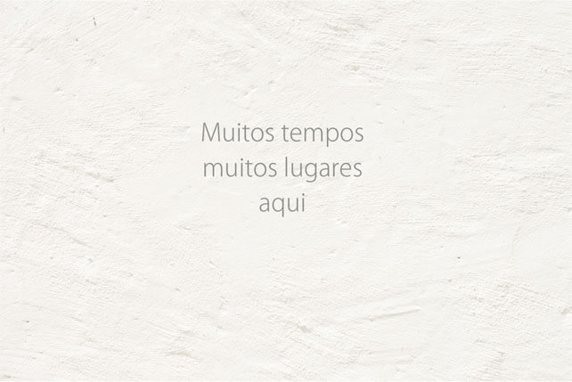 Imagem horizontal de uma parede branca com a sétima frase, em três linhas: Muitos tempos / muitos lugares / aqui.