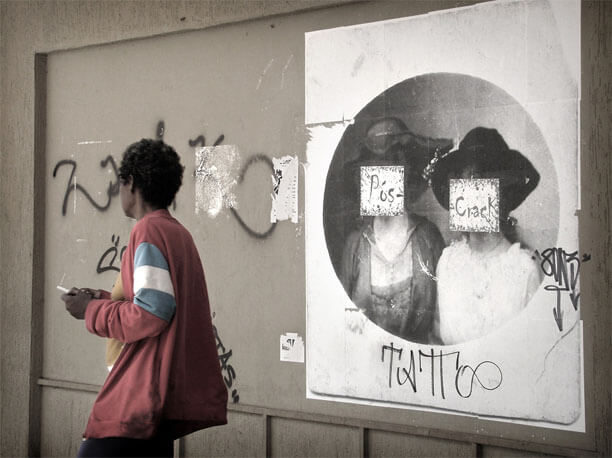 Foto no mesmo ângulo da anterior, um pouco mais perto da foto antiga, que aparece na metade direita. A margem branca da foto antiga tem dois nomes pixados com caneta grossa. Sobre o rosto de cada mulher está colado um papel A4; no primeiro está escrito "pós-" e no segundo "crack". Há uma pessoa em movimento no lado esquerdo da foto maior, vista das coxas para cima. Tem cabelo curto, está de lado/costas e segura um cigarro apagado nas mãos, que estão juntas na frente do corpo. Usa calça e um moletom vermelho grande demais, velho e de aparência suja.