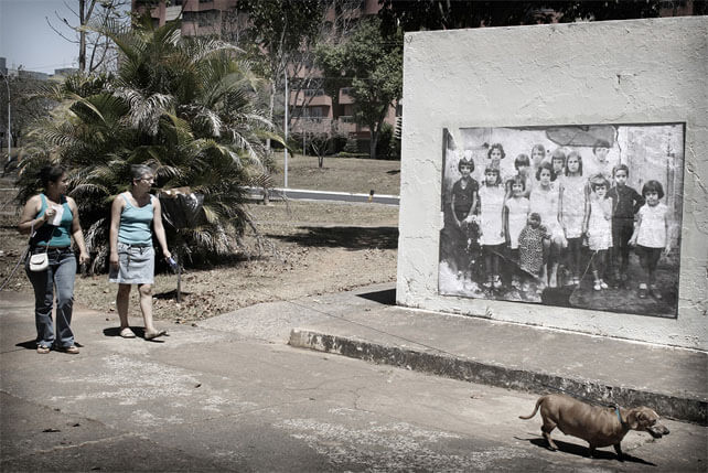 Foto horizontal de duas mulheres passeando com um cachorro e olhando uma foto antiga colada na parede. A foto colada tem cerca de 3m x 2m e mostra um grupo de 14 crianças (é a mesma da imagem anterior). As duas mulheres estão no canto esquerdo da foto e o cachorro no canto direito, já quase saindo do quadro. Atrás das mulheres vê-se uma grande planta, árvores e prédios de apartamentos ao fundo.