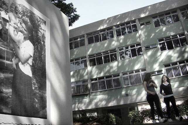 Foto horizontal enquadrada rente ao chão, mostrando, à esquerda, uma parede branca com uma grande foto antiga. À direita, ocupando dois terços da imagem, há um prédio de três andares sobre pilotis, com fachada branca e verde clara. Duas adolescentes estão andando na direção da câmera, conversando, a cerca de quatro metros, no canto à direita. A foto antiga mostra uma mulher segurando um bebê, ambos sorrindo. Ela usa blusa clara e saia escura e comprida, e seu penteado parece ser dos anos 40. 