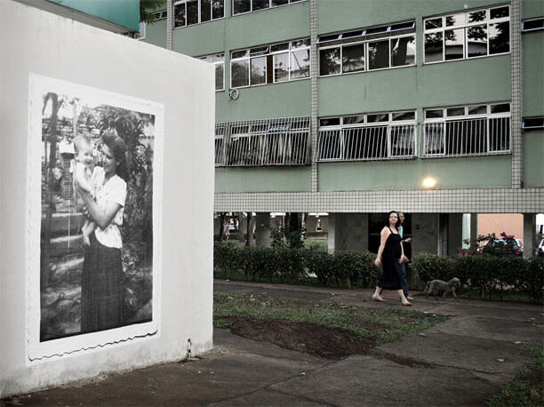 Foto horizontal do mesmo local, mostrando o mesmo ângulo entre a parede branca e o prédio, desta vez um pouco mais de perto e num enquadramento na altura normal. Na calçada em frente ao prédio, a cerca de oito metros de distância, está passando um casal. Eles estão de lado, andando para a direita e levando um cachorro pela guia. A mulher, que está mais próxima, olha para a foto na parede.