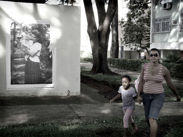 Foto horizontal da parede vista de frente, com a foto antiga à esquerda, a calçada e uma faixa de gramado na frente. Na parte direita vê-se o tronco de uma grande árvore, outra calçada e parte do prédio. Uma mulher de mãos dadas com uma menina vem em direção à câmera, já bem perto. A menina parece ter seis anos e está falando com a mulher.