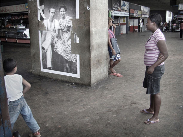 Foto horizontal mostrando a coluna com a foto antiga em perspectiva, na parte de cima, do centro para a esquerda da imagem. À direita há uma mulher de pé, de perfil, olhando a foto. Um pouco mais longe vê-se parte de uma pessoa de perfil, encostada atrás da coluna, olhando para longe. Mais perto da câmera, no cantinho da esquerda,está passando um menino de quatro ou cinco anos, já saindo do quadro. Ao fundo vê-se lojinhas da rodoviária, que está quase vazia.
