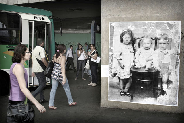 Foto horizontal mostrando, na metade da direita, uma coluna de concreto e uma grande foto antiga, quadrada. Na metade esquerda há um ônibus estacionado, três pessoas passando ao lado e outras ao fundo. Bem à esquerda e perto da câmera há uma mulher de perfil, andando em direção à foto antiga e olhando para ela com ar de espanto. A foto antiga mostra três crianças posando: uma menina de três anos, de pé; um bebê de seis meses sentado numa cadeira; e um menino de dois anos, de pé. A menina está à esquerda, com um braço atrás da cadeira, e o menino à direita, com uma mãozinha sobre o assento. O bebê está no meio, descalço, gordinho. A foto está muito maltratada, com parte das margens apagadas pelo tempo, várias manchas brancas e marcas de dobras. Algumas manchas estão sobre as crianças. Elas estão sérias, olhando para a frente. Há um certa melancolia nesse conjunto.