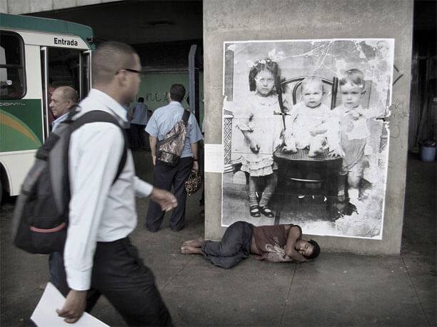 Foto horizontal do mesmo local, com o mesmo enquadramento. Junto à coluna com o retrato das crianças há um homem dormindo no chão, de lado, descalço. O lado de cima do seu corpo está sobre a margem inferior da foto antiga. Bem perto da câmera há um homem passando no lado esquerdo, indo para a direita e olhando a foto. Atrás dele vê-se parte de um ônibus, dois homens com camisa de uniforme azul claro e pessoas no fundo, ao longe.