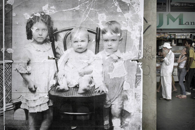 Foto horizontal mostrando um detalhe da foto antiga, que ocupa quase toda a imagem. Os rostos das crianças estão bem visíveis, e pode-se ver seus olhares sérios e intensos. À direita há uma estreita faixa onde se vê o fundo, vários metros atrás: parte do balcão de uma lanchonete e três pessoas de perfil, viradas para a esquerda. A primeira é um velho de longas barbas brancas; chapéu branco de pano, com abas largas; camisa social branca, maior que seu número e amarrotada; e uma calça larga e branca. Ele está com a mão sobre a barba e olha para a frente, parecendo pensativo. A segunda é uma jovem de cabelos compridos e lisos, falando ao celular e inclinada para o lado, dando uma grande risada. A terceira é um jovem olhando para a frente, sério.