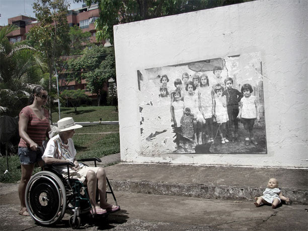 Foto horizontal de uma idosa numa cadeira de rodas, com vestido e chapéu brancos, empurrada por uma mulher jovem. Elas estão em meio perfil, no canto esquerdo. A idosa olha para uma boneca igual a um bebê, de vestidinho, deixada na calçada, no canto direito. Entre as mulheres e a boneca vê-se a parede branca com a mesma foto antiga das imagens anteriores, só que parte da imagem foi arrancada, no lado esquerdo e na parte de cima à direita. Atrás da parede há grama, plantas e um prédio.