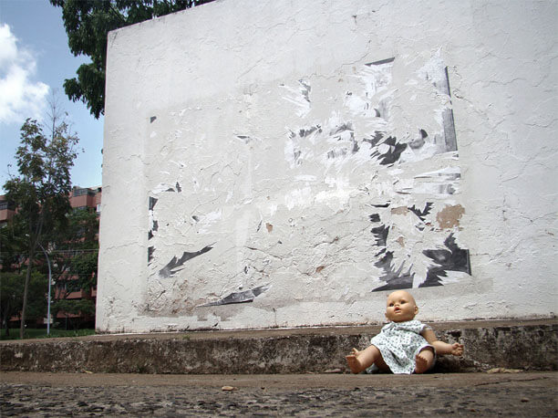 Foto horizontal de uma boneca igual a um bebê, careca e de vestidinho, sentada na calçada no canto direito. A imagem foi enquadrada de baixo, junto ao chão. Atrás da boneca vê-se a parede branca onde estava uma foto antiga, a mesma das imagens anteriores. A foto foi quase inteiramente arrancada e não se vê mais nenhuma criança. No lado esquerdo aparece uma estreita faixa do fundo, com árvores e um prédio.