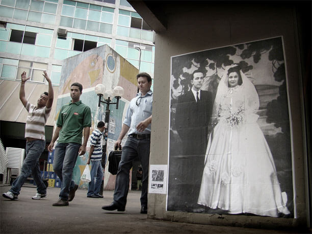 Foto horizontal de um pátio de cimento entre dois prédios. Na metade esquerda vê-se um prédio ao fundo, um homem olhando para trás e três homens passando de frente para a câmera. O da esquerda está fazendo um gesto com os braços para cima, em arco, como uma bailarina. O do meio olha para a câmera. Na metade direita está a parede do segundo prédio, onde foi colada uma foto antiga, em tamanho natural, de um casal de noivos. O noivo está de terno preto e a noiva de vestido branco longo, de cetim, com véu e buquê. Ele está sério; ela sorri levemente e é muito bonita. Ambos olham para a câmera.