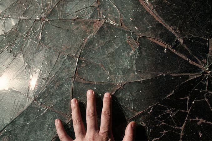 Foto horizontal; detalhe de um parabrisa de vidro rachado, verde escuro, com reflexos da luz do sol que entra pela esquerda. O lado direito está bem mais escuro e tem mais rachaduras. Na margem de baixo aparece metade de uma mão, com os dedos abertos pousados sobre o vidro, fazendo sombras escuras à direita.