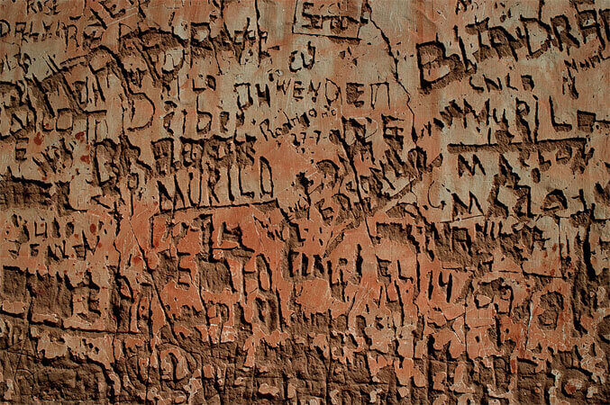 Foto horizontal; detalhe de uma parede marrom avermelhado, que pode ter sido branca ou amarela no passado, mas foi colorida pelo tempo e pelo barro. Toda a superfície está coberta por nomes de pessoas, que foram profundamente entalhados no reboco. Os nomes foram entalhados uns sobre os outros e muitos deles não são mais legíveis.