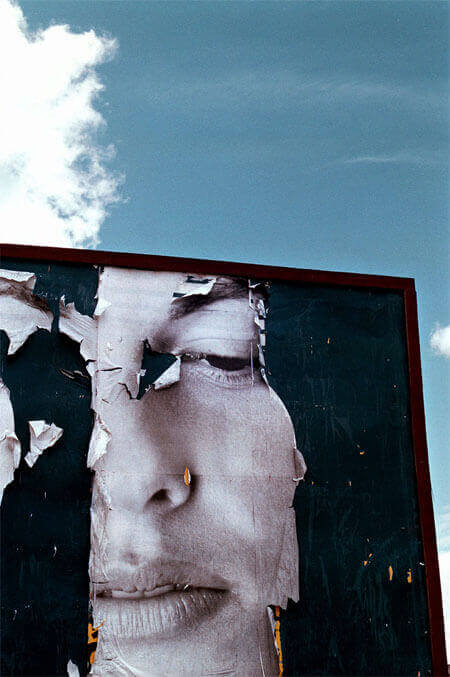 Foto vertical mostrando metade de um outdoor azul marinho, quase preto, que está rasgado numa larga faixa vertical. Nessa faixa vê-se o rosto de uma mulher em preto e branco. O rosto aparece pela metade: uma sobrancelha, um olho, parte do nariz e da boca. A mulher tem uma expressão melancólica e o olhar voltado para baixo. Há rasgos em algumas partes do seu rosto. Na metade de cima da foto está um céu azul e uma nuvem branca junto à margem esquerda.(Esta é a última imagem da série.)