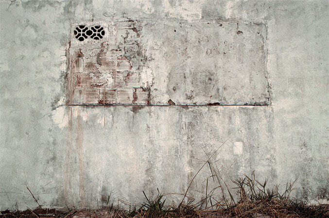 Foto horizontal; detalhe uma parede branca desgastada pelo tempo, que agora está cinza e escura, com muitas manchas. No centro da parede havia uma janela larga que foi toda tapada com tijolos, exceto por dois cobogós pequenos, no canto superior esquerdo. Essa janela tapada foi parcialmente rebocada e pintada de branço; vê-se vestígios dos tijolos vermelhos por baixo. No chão há uma faixa estreita de mato seco.