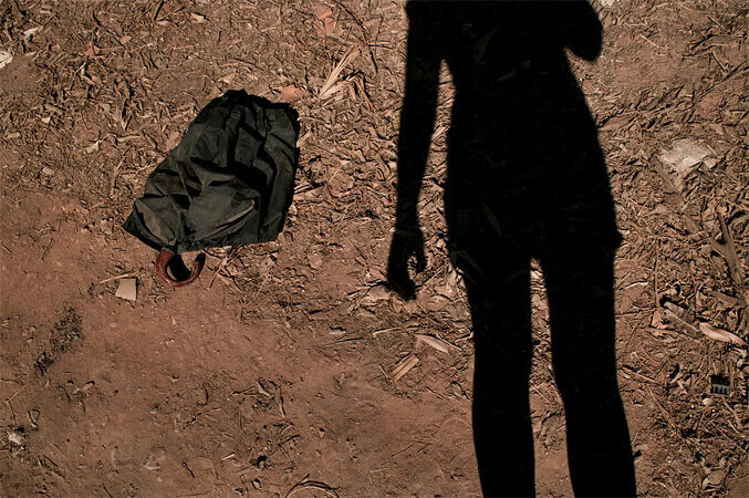 Foto horizontal da sombra de uma pessoa sobre um chão de terra marrom com folhas de mato seco, ao lado de um guarda-chuva preto parcialmente dobrado. O guarda-chuva aparece no lado esquerdo e a sombra no direito, cortada na altura das canelas e dos ombros. Está ligeiramente inclinada em direção ao guarda-chuva; um dos braços está esticado, um pouco afastado, e o outro está recolhido junto ao corpo.