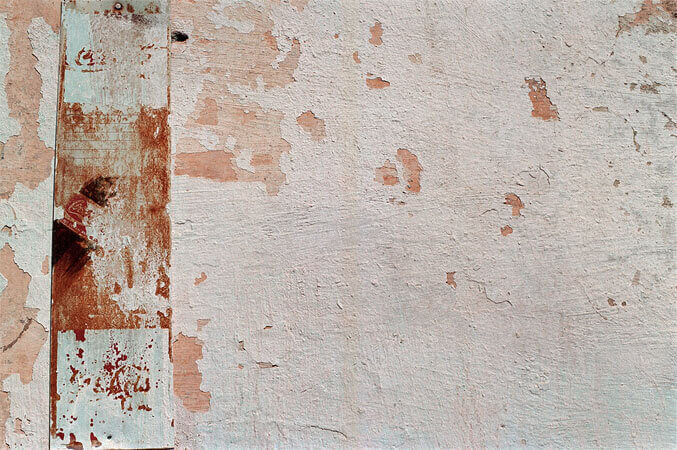 Foto horizontal; detalhe de uma parede branca com a pintura descascada, deixando ver a pintura anterior, avermelhada. O branco da parede tem manchas cinza-azuladas. O lado esquerdo da foto é atravessado por uma estreita faixa de metal na vertical, que parece ter sido um antigo anúncio de coca cola; está muito enferrujado e tem tons de marrom, vermelho e branco. As palavras foram todas apagadas pelo tempo.
