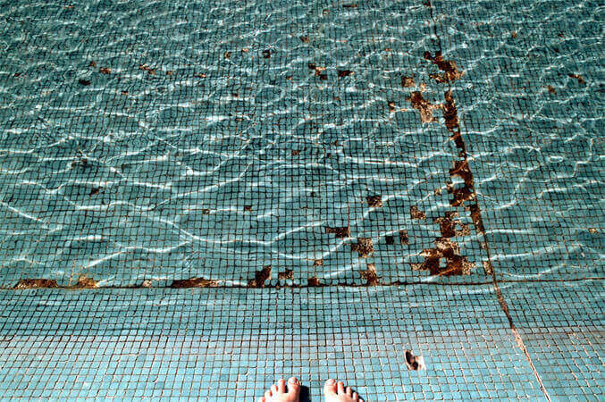 Foto horizontal de um espelho d'água revestido de pequenas pastilhas azul-turquesa. Faltam muitas pastilhas, e isso cria manchas marrons no fundo. A água é transparente, com desenhos de pequenas ondas feitos pela luz do sol. Na parte seca, junto à margem de baixo, aparece a metade de dois pés descalços. 