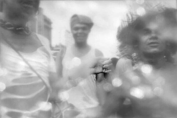 Foto horizontal de três homens de frente para a câmera, da cintura para cima. O da direita está sem camisa, usa um chapéu de palha com a aba desfiada e encara a câmera. Está cortado na margem direita. O do centro está dois passos atrás, usa um pano na cabeça e sorri para a câmera. O da esquerda está cortado na margem e aparece da boca para baixo. Usa brinco, batom, uma grossa corrente no pescoço e uma blusa com grandes buracos mostrando a barriga. Sua a mão está estendida para tocar o homem ao lado. A imagem parece ter um anteparo na lente que cria várias formas brancas, pequenas e arredondadas. Esse anteparo deixa tudo desfocado, exceto por um pequeno buraco onde se vê melhor parte da mão estendida.