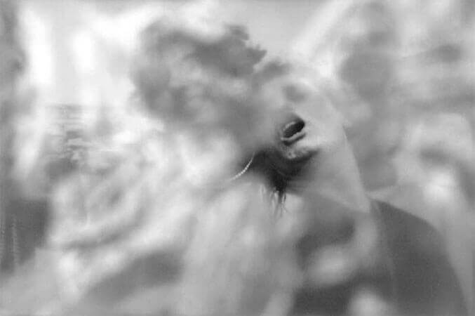 Foto horizontal de três homens vestidos de mulher, com perucas e maquiagem. Eles aparecem do peito para cima. O do meio está com a boca aberta e a cabeça jogada para trás, numa pose sensual caricata. O anteparo na lente deixa tudo muito desfocado e coberto por manchas brancas e compridas. Há um buraco no anteparo onde se vê com clareza a boca e queixo do homem do meio.