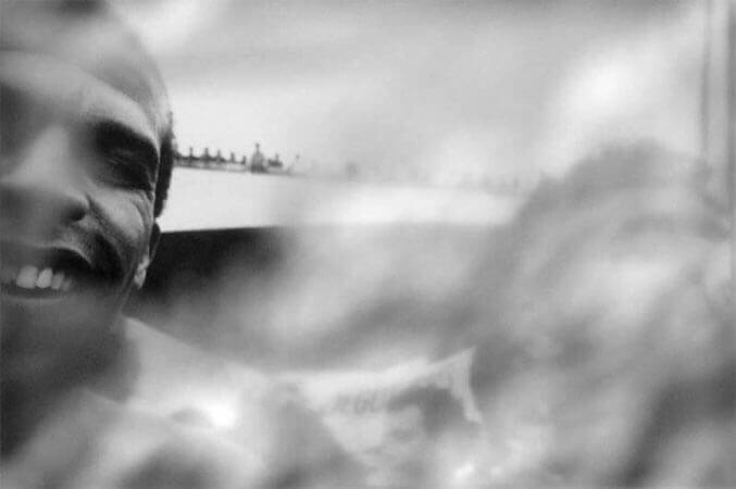 Foto horizontal mostrando ao longe um viaduto com um grupo de pessoas no guarda-corpo, vendo o bloco passar. Em primeiro plano há o rosto de dois homens bem perto da câmera, ambos cortados nas margens laterais. O da direita é quase um borrão desfocado pelo anteparo na lente e parece estar bebendo de uma latinha. O da esquerda está dando um grande sorriso pra a câmera. Há um buraco de bom tamanho no anteparo, que permite ver bem seu sorriso e seu olho direito.