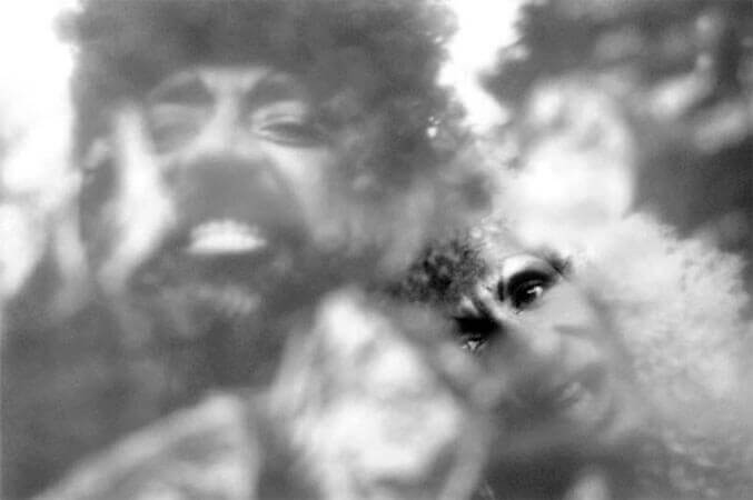 Foto horizontal do rosto de dois homens com perucas compridas, olhando para a câmera. O da esquerda é barbudo e sua peruca é escura e cacheada. Ele arreganha os dentes num sorriso grande e forçado. O da direita parece estar com a cabeça deitada no seu ombro, e sua peruca é clara e cacheada. Seu olhar é fixo e intenso, e ele está com a boca aberta, como quem está falando. O anteparo na lente deixa tudo desfocado, menos uma pequena faixa onde se vê nitidamente os olhos do segundo homem.