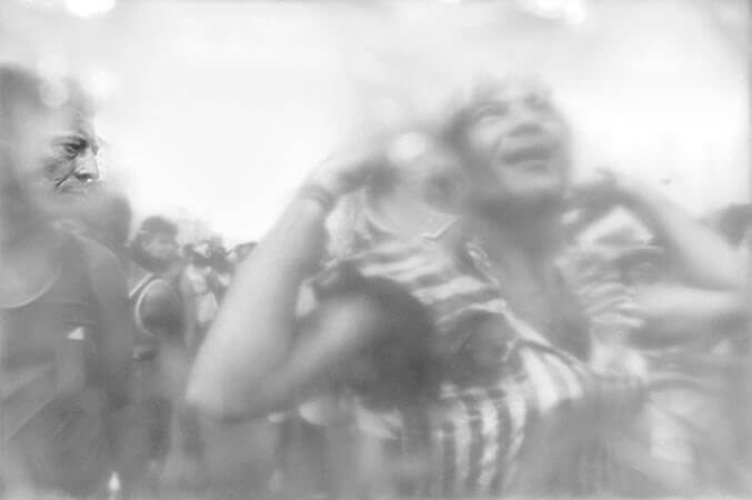 Foto horizontal mostrando duas pessoas em primeiro plano e um bloco de carnaval atrás. O homem da direita ocupa quase toda a imagem e aparece da barriga para cima. Está de peruca lisa com franja e usa uma blusa ou vestido brilhante e listrado. Ele sorri olhando para cima e levanta as duas mãos como quem vai ajeitar o cabelo, num gesto caricato, com os cotovelos bem abertos. Cortado na margem esquerda da foto há um homem de perfil, de roupa comum, olhando para o gesto do outro com o cenho franzido e uma expressão de reprovação. O anteparo na lente deixa tudo desfocado, menos uma pequena faixa onde se vê nitidamente o nariz e os olhos do homem sério.