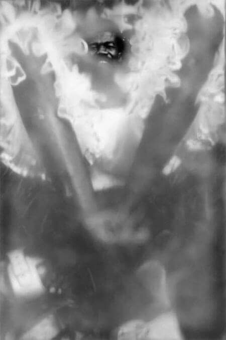 Foto vertical de um homem deitado na grama, com enquadramento bem próximo, rente ao chão. Ele tem pernas compridas e finas que fazem um V ocupando quase toda a imagem. Seus pés estão cruzados e de tamancos brancos. As pernas estão emolduradas por uma fantasia branca de tule, com muitos babados. O rosto do homem está levantado, olhando para a câmera e sorrindo. Sua face parece pequena por causa da perspectiva. O anteparo na lente deixa tudo desfocado, menos uma pequena área onde se vê o rosto.(Esta é a última imagem da série.)