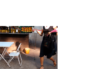 Foto horizontal de uma mulher passando apressada na rua, cortada dos ombros para cima. Ela usa vestido preto sobre uma blusa branca de mangas curtas, calça sandálias pretas e o pé que está na frente está cortado pela margem de baixo da foto. Está entrando na imagem pela margem direita, e carrega várias coisas nas duas mãos (celular, bolsa e casaco pretos, duas pastas rosas). O chão é um pátio de cimento. Na metade esquerda da foto há um quiosque de frutas, visto do chão até o balcão, onde estão expostos mamões e bananas. Na frente do quiosque aparece metade de uma mesinha branca de metal com uma cadeira. Bem no centro da imagem há um cesto de lixo laranja vivo, iluminado pela luz do sol do começo da manhã. A luz bate em outras pequenas partes da foto.