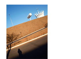 Foto vertical com enquadramento bastante inclinado, pendendo para a esquerda. No terço de cima vê-se um céu azul sem nuvens, e parte de três elementos, da direita para a esquerda: o topo da catedral de Brasília; a torre dos sinos; e um poste branco e curvo. No terço central há um barranco de terra coberto por grama seca, em tons de marrom avermelhado. No terço de baixo vê-se, da esquerda para a direita: parte de uma rua marrom com meio-fio branco; a sombra de uma grande árvore que sai do canto esquerdo; a sombra de uma pessoa que parece estar tirando a foto; e uma grande sombra triangular, que parte do canto esquerdo e sobe para a direita.