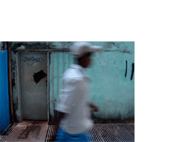 Foto horizontal de um homem passando de perfil, borrado, no centro da imagem, do quadril para cima. Ele usa calça azul, camisa branca e boné cinza clarinho. Atrás dele há uma porta cinza com uma grande mancha preta de ferrugem e um nome pixado. Ela está numa parede verde clara com pintura muito velha, manchas pretas de umidade e algumas pixações. Numa estreita faixa à esquerda a parede está pintada de azul novo e vivo, no mesmo tom da calça do homem. No chão há uma grade de ventilação enferrujada, acompanhando a linha da parede.