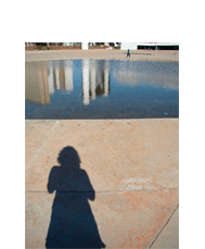 Foto vertical de um imenso pátio de concreto bege com um espelho d'água na parte de cima, cortado nas margens. Na parte de baixo há a sombra grande de uma mulher que parece estar tirando a foto. A sombra é longa e está do centro para a esquerda. O espelho d'água reflete o céu azul escuro e o topo de prédios altos, em tons de preto, branco e bege. Do outro lado dele há um homem passando, bem ao longe, e ainda mais ao longe três pessoas quase indistinguíveis, passando onde termina o pátio, junto ao topo da foto.