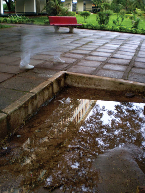 Foto vertical de um pátio de cimento quadriculado onde se vê dois pés com sapatos brancos e duas pernas com calça branca, dando uma larga passada. Estão no centro/esquerda, a meia distância. Parecem ser de um fantasma, pois não se vê o resto das pernas: da barra da calça para cima a imagem vai ficando borrada e transparente até sumir. Ao longe há um banco vermelho e um gramado verde, tudo em cores muito vivas. O chão está molhado. Da metade da foto para baixo vê-se um meio-fio branco formando uma quina, apontando para o pé do fantasma. O meio-fio delimita um espaço de areia com uma grande poça d'água, que reflete a copa de uma árvore e um prédio comprido e baixo.