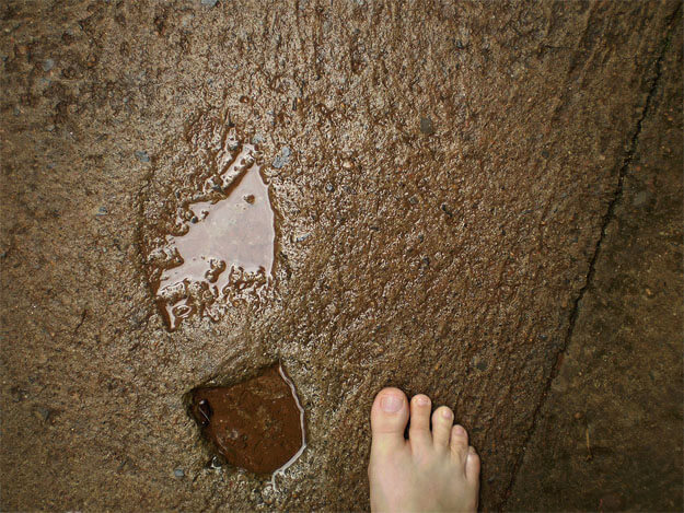 Foto horizontal mostrando o detalhe de uma calçada de cimento com uma pegada antiga, feita por um sapato ou bota quando o cimento estava fresco. A calçada está molhada e a pegada está cheia de água. Ela aparede do meio para a esquerda da foto, e à sua direita há metade de um pé descalço.
