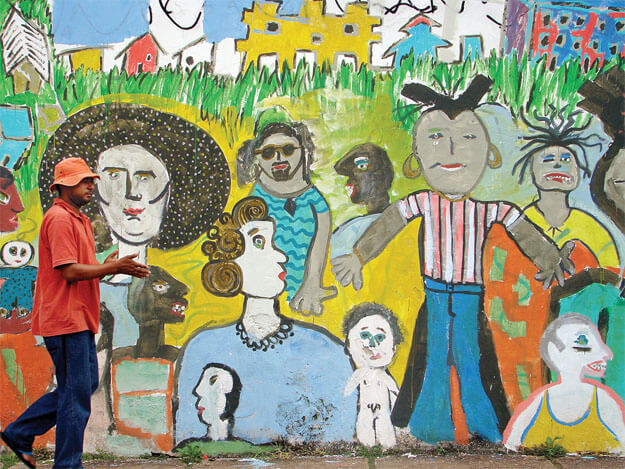 Foto horizontal mostrando o detalhe de uma parede vista de frente. Junto à margem esquerda está passando um homem de perfil, com calça azul, camisa laranja e chapéu de pano laranja. Ele faz um gesto com as duas mãos, como quem fala sozinho. A parede junto a ele está toda coberta uma pintura de cores vivas, representando uma dúzia de pessoas de idades e estilos diferentes, de frente e de perfil.