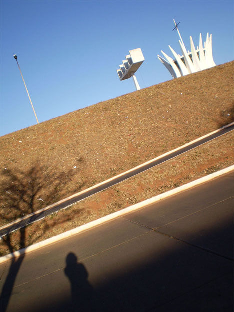 Foto vertical com enquadramento bastante inclinado, pendendo para a esquerda. No terço de cima vê-se um céu azul sem nuvens, e parte de três elementos, da direita para a esquerda: o topo da catedral de Brasília; a torre dos sinos; e um poste branco e curvo. No terço central há um barranco de terra coberto por grama seca, em tons de marrom avermelhado. No terço de baixo vê-se, da esquerda para a direita: parte de uma rua marrom com meio-fio branco; a sombra de uma grande árvore que sai do canto esquerdo; a sombra de uma pessoa que parece estar tirando a foto; e uma grande sombra triangular, que parte do canto esquerdo e sobe para a direita.