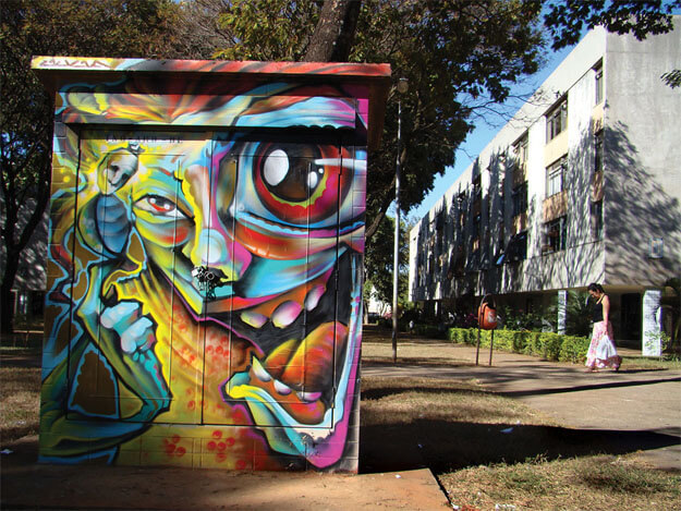 Foto horizontal mostrando em primeiro plano um retângulo de concreto inteiramente grafitado por um artista. Ele ocupa a metade esquerda da foto e mostra uma figura grande, meio gente e meio fantástica, com um olho pequeno e outro enorme. Ela ri com a boca bem aberta e segura algo que parece um pequeno besouro com uma cabeça de boneco de pano. A pintura tem cores fortes e vibrantes. Na metade direita da foto vê-se uma calçada em perpectiva em direção ao fundo, acompanhando um prédio comprido de três andares, e várias árvores. Há uma mulher passando ao longe, de cabeça baixa, carregando duas sacolas de plástico.