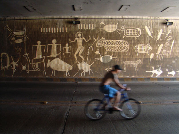 Foto horizontal mostrando o teto de um viaduto, o chão de concreto e, ao fundo, um paredão com cinco ou seis metros de altura. O paredão é revestido de pequenos ladrilhos compridos e está marrom escuro de sujeira, quase preto. Nele há grandes desenhos brancos feitos com arte reversa (limpando em vez de pintar). Os desenhos imitam arte rupestre, com imagens de animais e figuras humanas. A meia distância, do centro para a margem direita da foto, há um homem numa bicicleta azul, de bermuda azul, sem camisa e de boné preto. Ele vai em direção à direita e sua imagem está borrada pela velocidade.