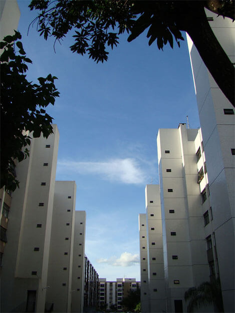 Foto vertical de dois prédios brancos em perspectiva, de seis andares e bem compridos. Eles estão cortados pelas duas margens laterais e formam um triângulo que emoldura o céu azul, com poucas nuvens. Há outro prédio ao fundo, bem ao longe, "fechando" a ponta do triângulo. Em primeiro plano vê-se parte da copa de duas árvores, uma em cada margem superior. Elas formam uma mancha preta, pois estão na sombra.