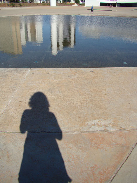Foto vertical de um imenso pátio de concreto bege com um espelho d'água na parte de cima, cortado nas margens. Na parte de baixo há a sombra grande de uma mulher que parece estar tirando a foto. A sombra é longa e está do centro para a esquerda. O espelho d'água reflete o céu azul escuro e o topo de prédios altos, em tons de preto, branco e bege. Do outro lado dele há um homem passando, bem ao longe, e ainda mais ao longe três pessoas quase indistinguíveis, passando onde termina o pátio, junto ao topo da foto.
